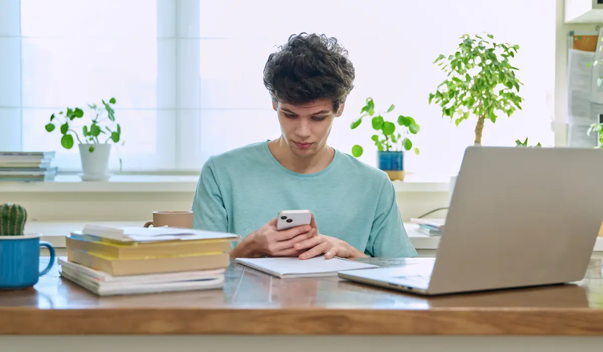 Teen using a phone while studying at a desk with a laptop and books, showing posture habits linked to teen neck pain.