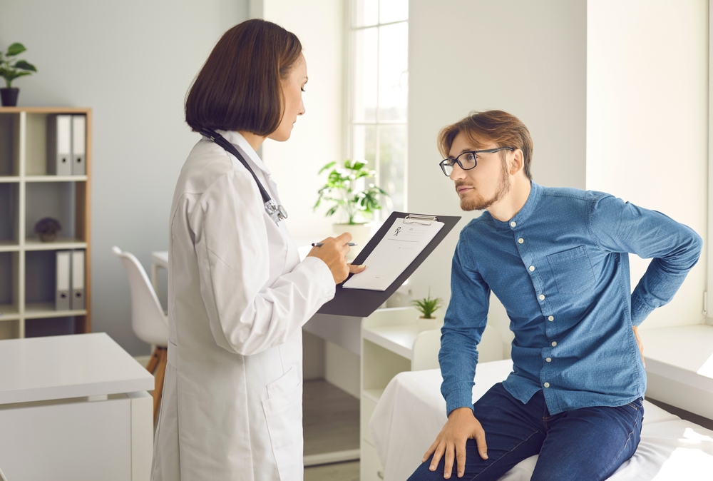 A female doctor in a white coat with a stethoscope holding a clipboard and talking to a male patient who is sitting on a treatment table and holding his lower back in pain.
