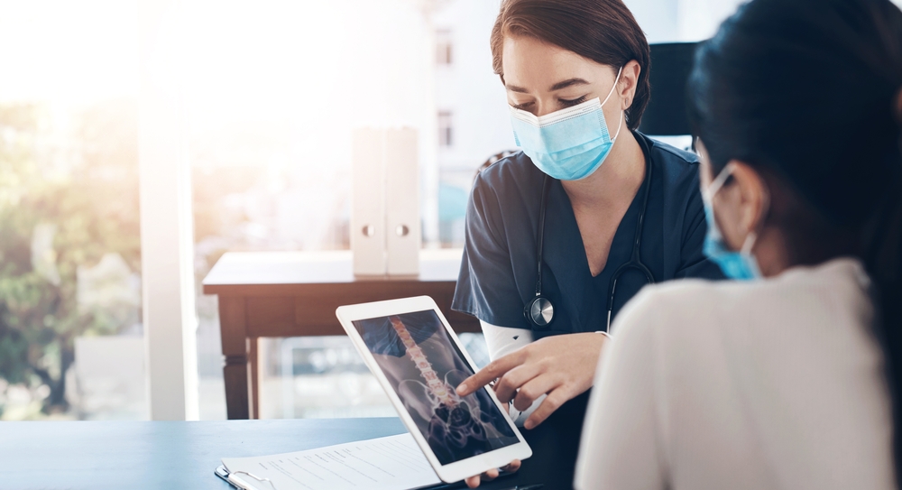 A doctor in a blue mask pointing at a lumbar spinal X-ray on a tablet while consulting with a patient.