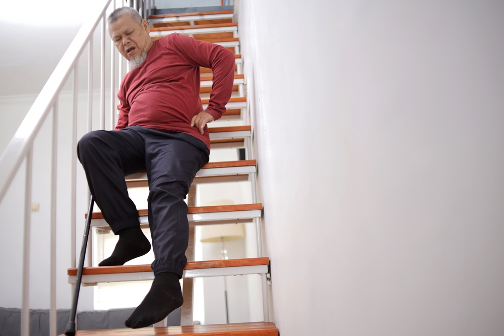 An elderly man with a cane sitting on a staircase holding his lower back with a pained facial expression.