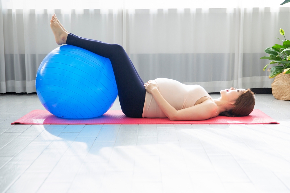 A pregnant woman lying on a yoga mat with her legs elevated on a blue stability ball to relieve lower back pressure.
