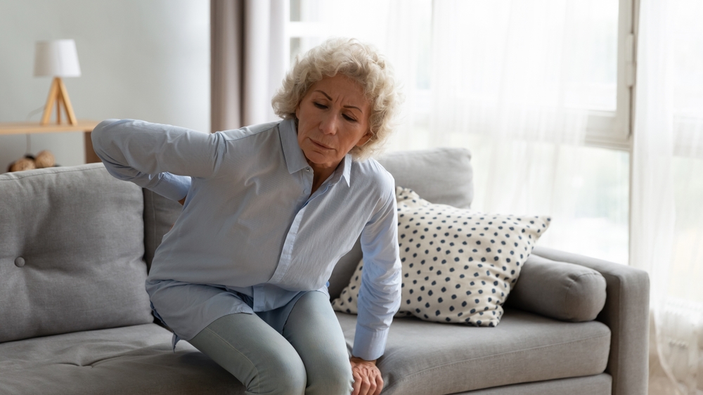 Senior woman holding her lower back in pain while attempting to stand up from a grey sofa in a brightly lit living room.