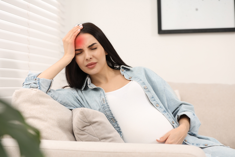 A pregnant woman sitting on a couch holding her forehead in pain with a red glow indicating a headache.