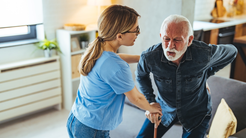 A young female caregiver assisting a senior man with a cane as he struggles with lower back pain in a living room.