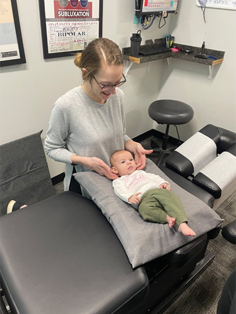 A pediatric chiropractor gently performing a manual cranial and spinal assessment on an infant in a onesie and green pants on a specialized chiropractic table.