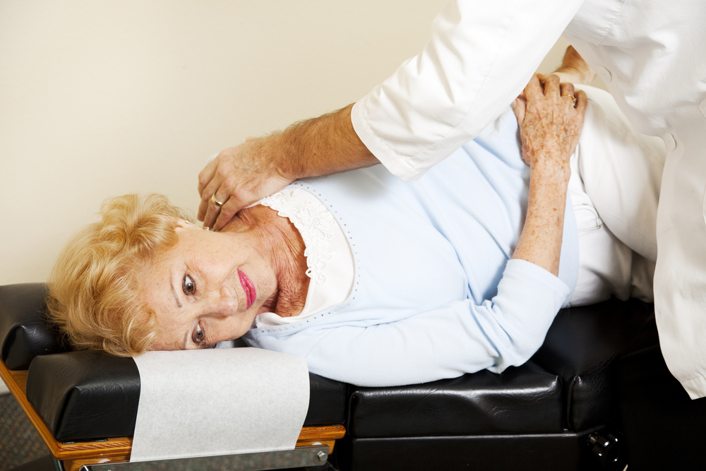 A senior woman with blonde hair lying on a black chiropractic adjustment table while a practitioner in a white lab coat performs a neck and shoulder realignment.