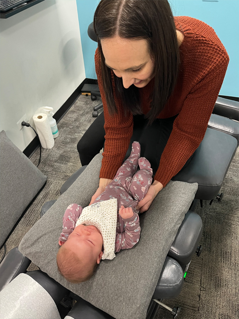 A pediatric chiropractor gently performing a manual assessment on an infant in a patterned floral onesie and bib on a black chiropractic table.
