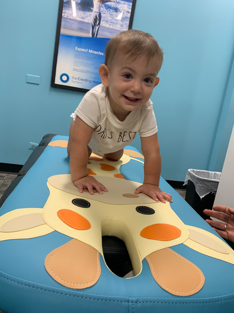 A happy toddler crawling on a blue pediatric chiropractic table designed with a large giraffe face, smiling toward the camera during a well-child visit.