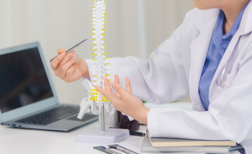 Close-up of a female doctor in a white lab coat using a pen to point at a human spine model during a clinical consultation.