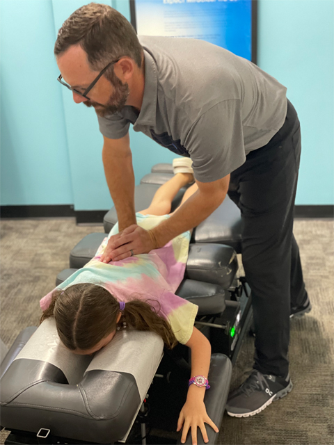 A pediatric chiropractor gently performing a manual spinal alignment or assessment on a young child in a tie-dye shirt on a specialized table.